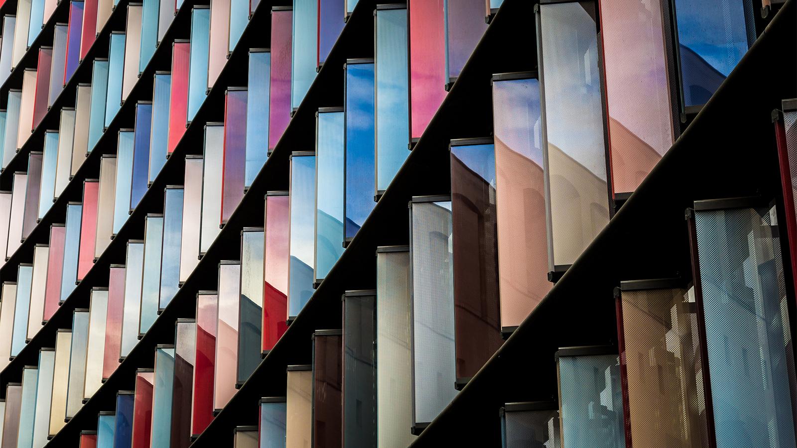 Abstract, close-up stock photograph looking up at contemporary architecture in London, UK. The office building exterior is composed of coloured glass panels, which reflect the blue sky and cloudscape above.